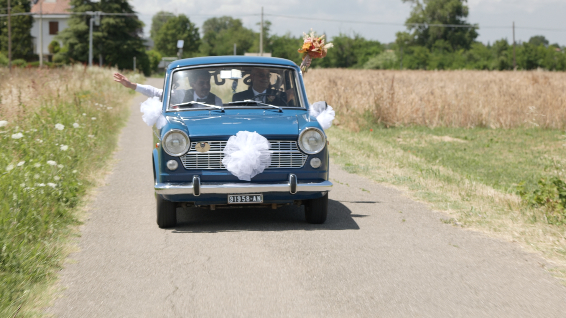 Fiat vintage blu decorata con fiocchi di tulle bianco su una strada di campagna, un braccio con il bouquet fuori dal finestrino